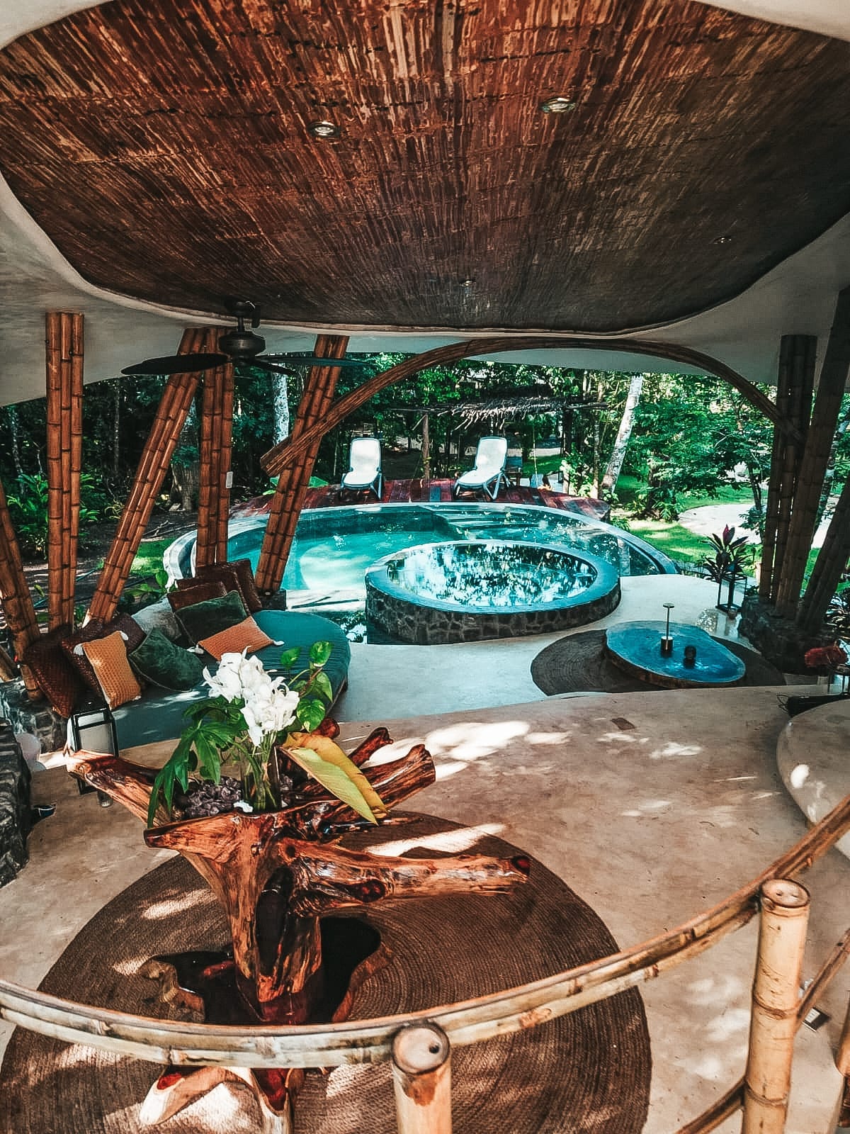 A spacious living area surrounded by natural bamboo features leads to an outdoor pool and jacuzzi area. Comfortable seating arrangements are present alongside lush greenery, while a wooden table adorned with a floral centerpiece adds a touch of nature indoors.