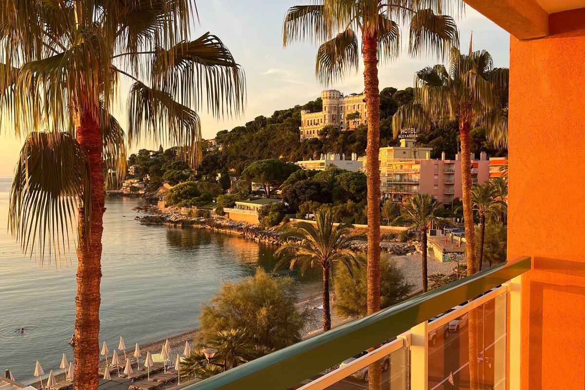 A balcony view reveals a tranquil beach scene framed by tall palm trees. The sun sets, casting warm hues over the landscape, with gentle waves lapping against the shore. Loungers are positioned along the shoreline, and lush greenery lines the background.