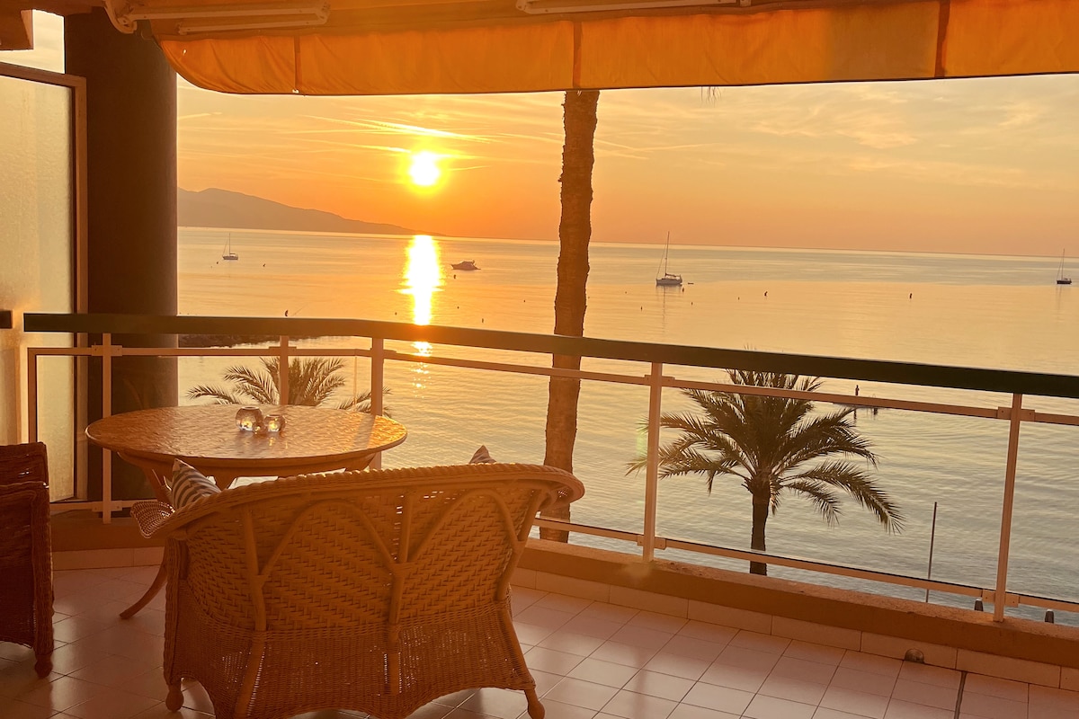 A balcony setting features a round table with woven seating, overlooking a serene sunset on the sea. The sun casts a warm glow on the water, and palm trees are visible along the shoreline, creating a tranquil atmosphere.