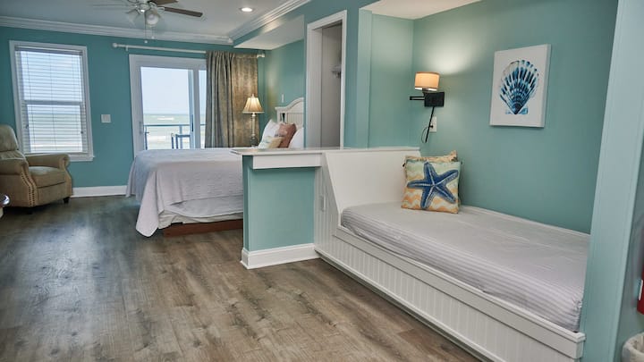 Pohľad na izbu vrátane dennej postele/sedacej časti, manželskej postele, pohodlného kresla, zadného okna a balkóna s výhľadom na oceán.