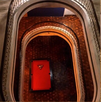 A view from above showcases a circular staircase with elegant railings. At the center, a vibrant red pool table is positioned, surrounded by a textured floor. The warm lighting highlights the architectural curves of the staircase and adds depth to the space.
