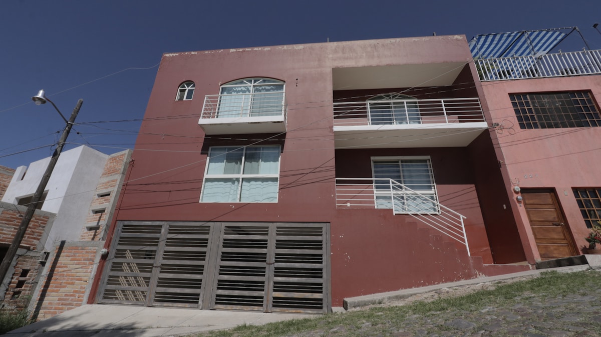 An exterior view of a two-story building with a red facade. Large windows are situated on both levels, allowing for ample natural light. A wooden gate is visible at the front, accompanied by a staircase leading to the entrance. The surrounding area features other residential structures.