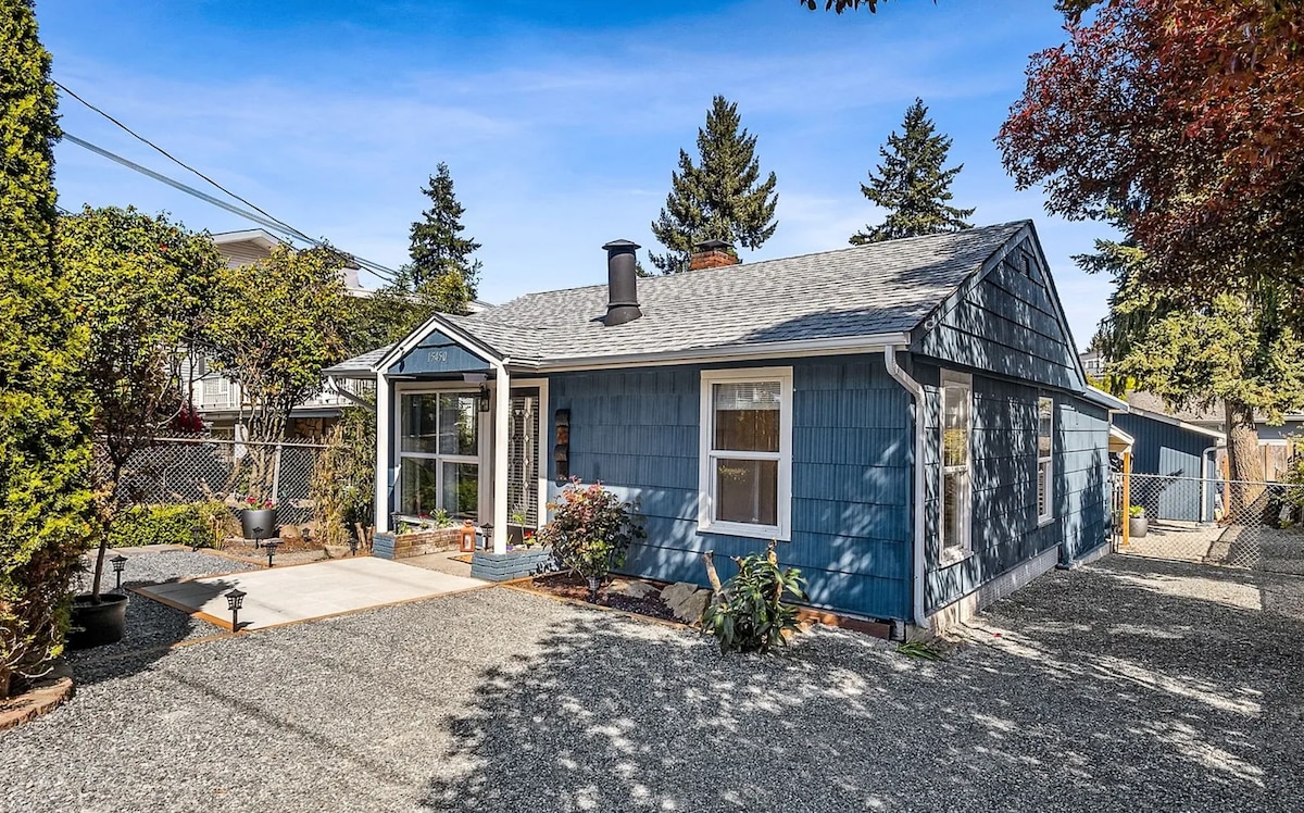 A cozy blue home with a gravel driveway is framed by greenery. Large glass doors provide natural light and a view of the covered entry. A welcoming outdoor area is suggested by the surrounding plants and trees.