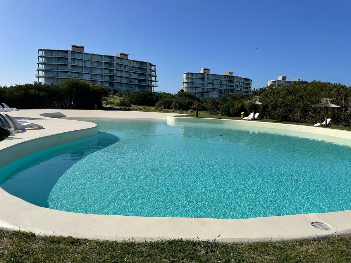 A tranquil outdoor swimming pool is depicted, featuring clear turquoise water with a gently sloping edge. In the background, modern buildings rise against a clear blue sky, while shaded lounge chairs are positioned along the grassy area nearby.