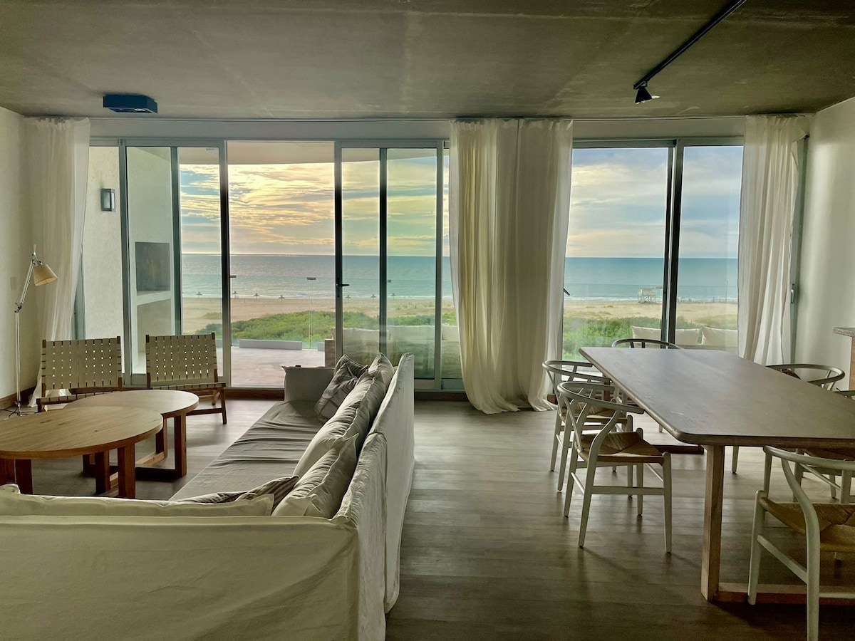 A spacious living area is presented, featuring large glass doors that provide uninterrupted views of the ocean. Soft curtains frame the windows, allowing natural light to enhance the simple, neutral decor. A round wooden table and a rectangular dining table are visible, along with comfortable seating.
