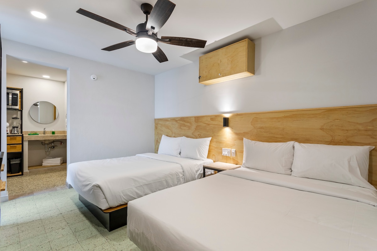 A well-appointed guest room features two spacious beds with white linens against a warm wood-paneled wall. A ceiling fan provides air circulation, while soft lighting creates an inviting ambiance. A mirrored vanity area with toiletries is visible in the background, enhancing convenience for guests.