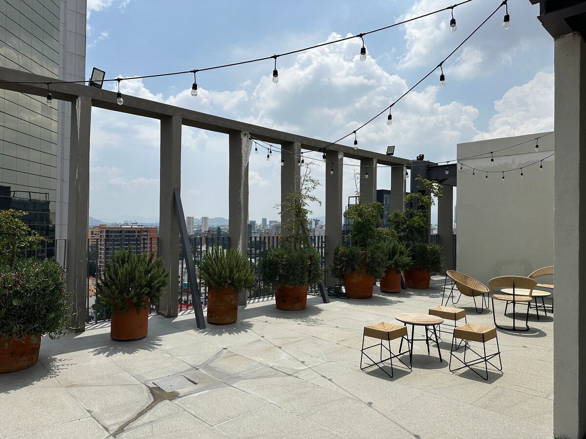 A private terrace is presented, featuring several potted plants and comfortable seating arrangements. String lights are hung overhead, providing a charming ambiance. The city skyline is visible in the background, enhancing the outdoor space's integration with the urban setting.