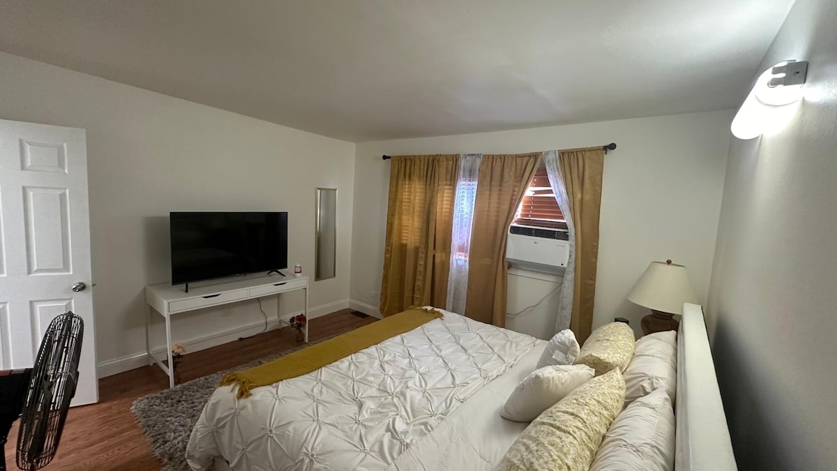 A spacious bedroom is arranged with a king-size bed, complemented by decorative pillows and a textured throw. A flat-screen TV is mounted on the wall opposite the bed, while a window-unit air conditioner adds comfort. Light drapes frame the window, allowing natural light to enter.