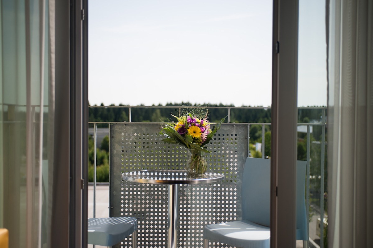 A small balcony is viewed through glass doors, featuring a round table paired with two light blue chairs. A colorful bouquet of flowers sits at the center of the table. Green foliage is visible in the background under a clear sky.