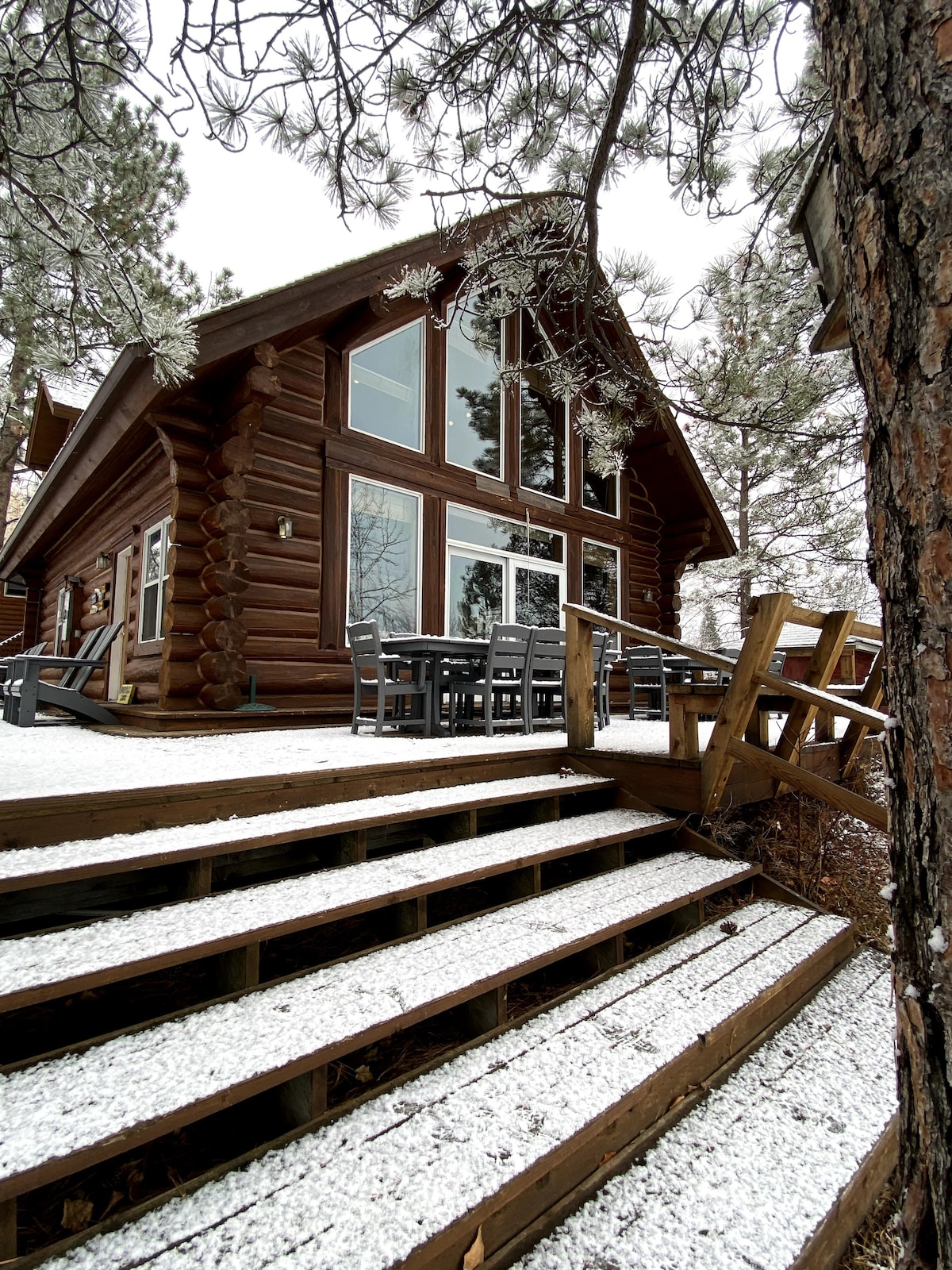 The exterior of a log cabin is depicted, featuring large windows that allow natural light to fill the interior. A set of steps leads up to a wooden deck, where several outdoor chairs are arranged for seating. A light layer of snow covers the ground.