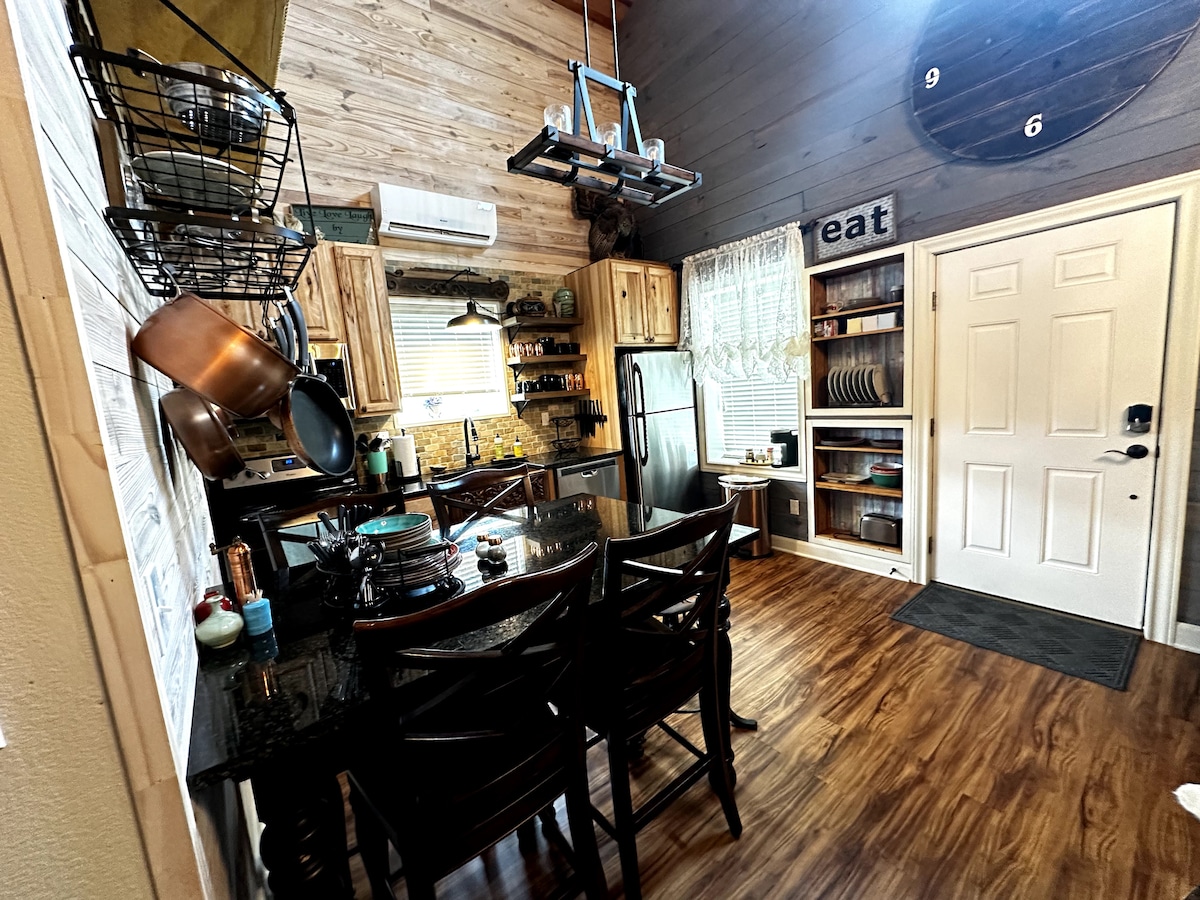 A well-equipped kitchen is visible, featuring dark cabinets and a granite-top island with seating for four. Various cooking utensils and pots hang above the island. Natural light enters from a nearby window, illuminating the space, and a door leads to the outside.