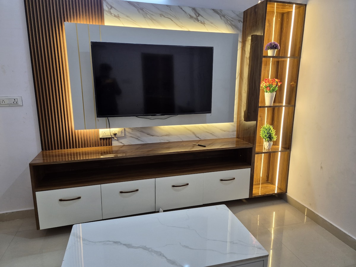 A modern living area features a flat-screen television mounted on a marble-patterned wall. Below, a wooden console with three drawers offers storage. A tall cabinet with illuminated shelves displays decorative flowers, complementing the sleek coffee table set on a polished floor.
