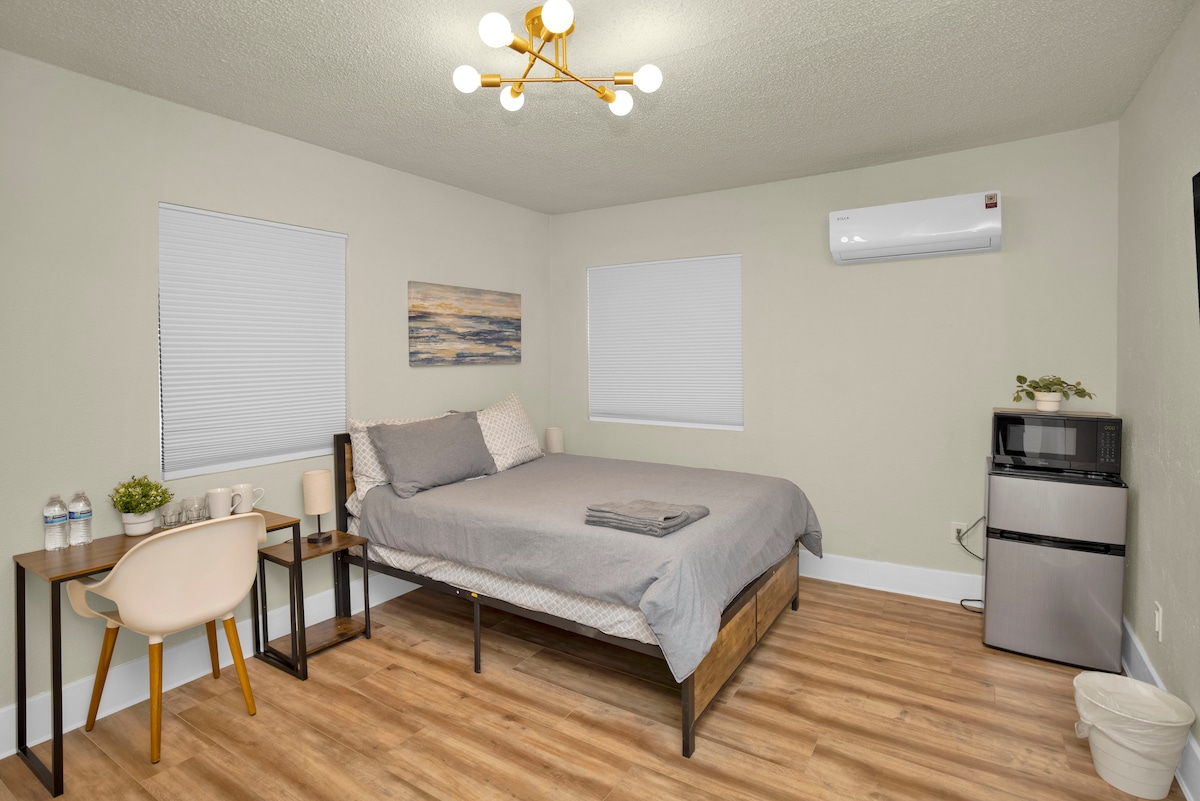 The room features a spacious bed covered with a grey blanket, complemented by light-coloured walls and a hardwood vinyl floor. Two windows allow natural light to fill the space. A desk with two water bottles and a mini fridge are also visible.