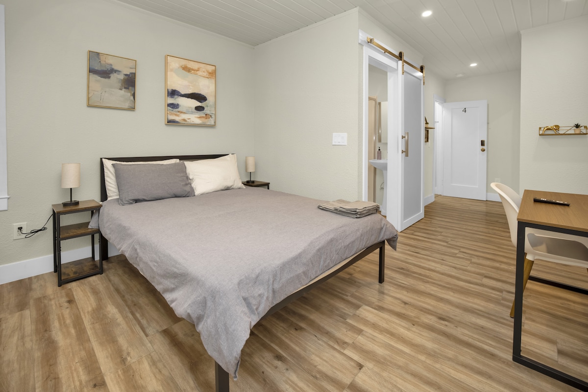 A well-lit guest suite features hardwood vinyl flooring and minimalist decor. A spacious bed is dressed in light bedding, complemented by bedside lamps on either side. A small desk with a chair is positioned nearby, and sliding doors provide access to the private bathroom.