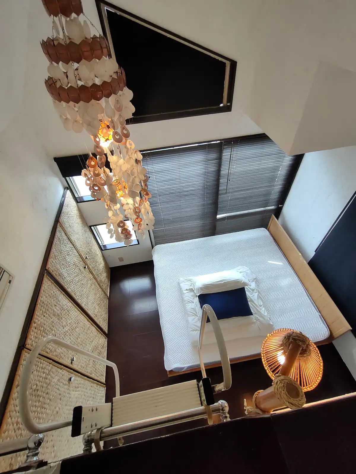 An overhead view of the bedroom is presented, showcasing a spacious bed with a dark blue accent pillow. Soft, natural light filters through the windows, which are dressed in dark blinds. A unique chandelier adds visual interest above, while textured wall panels provide character.