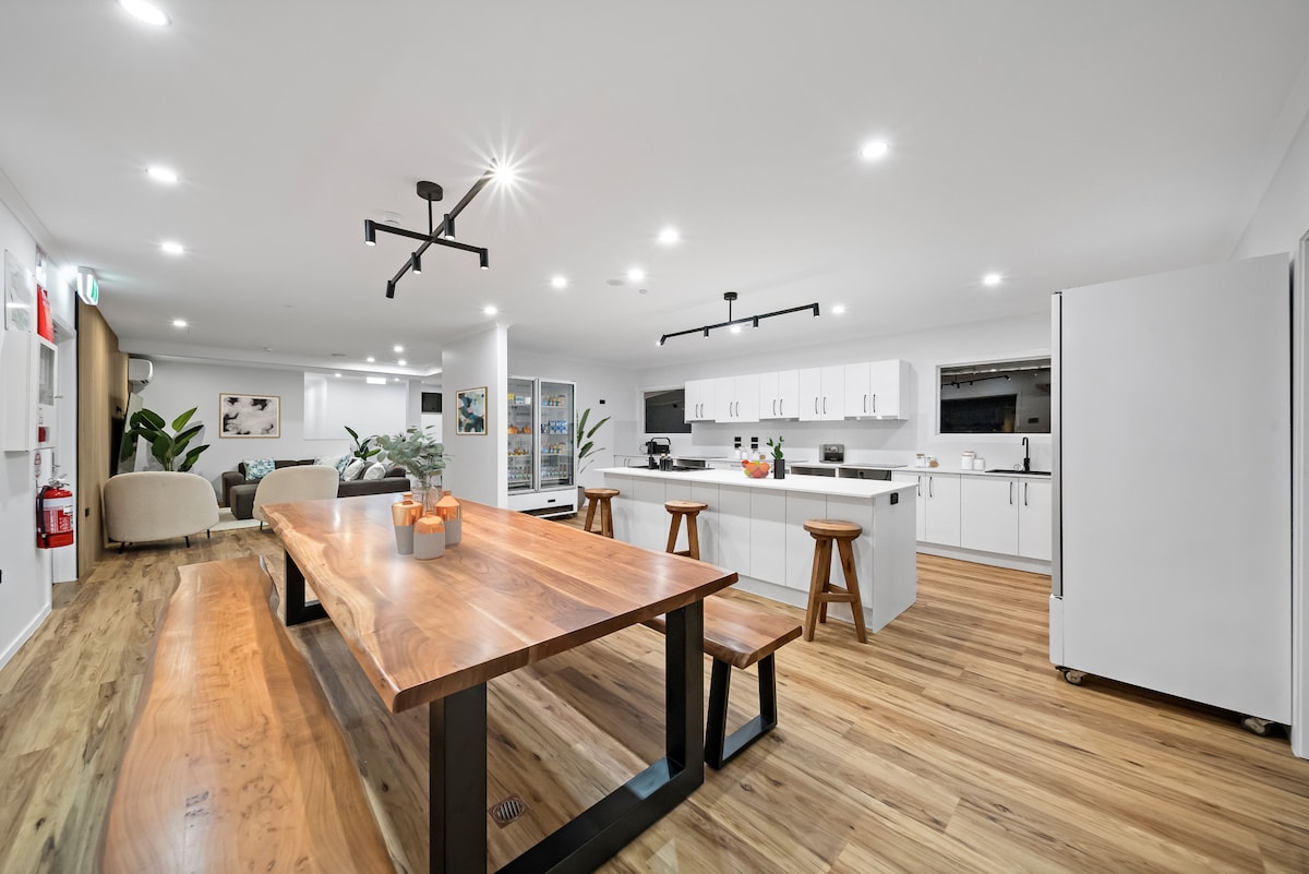 A spacious kitchen and dining area features modern appliances and a large wooden dining table accompanied by three stools. The bright space is complemented by warm lighting and touches of greenery, creating a functional yet inviting atmosphere for cooking and dining.