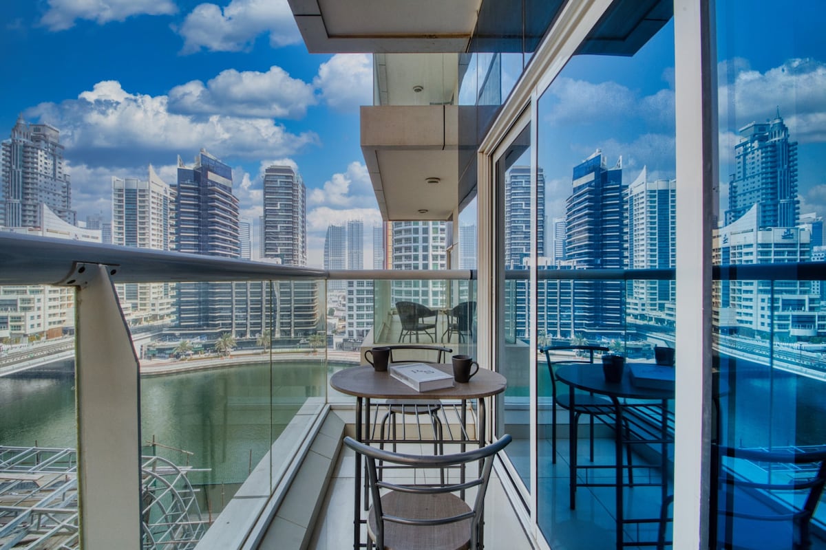 A balcony with views of the Dubai Marina is presented, featuring a small round dining table and two chairs. The glass railing offers unobstructed sights of the skyscrapers and waterway, while the blue sky with scattered clouds enhances the scenic backdrop.