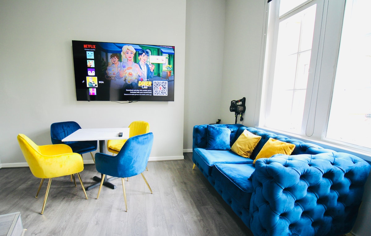 A modern living area features a plush blue tufted sofa complemented by yellow accent chairs. A small white dining table is positioned nearby. A flat-screen TV is mounted on the wall, displaying content, while natural light filters through large windows.