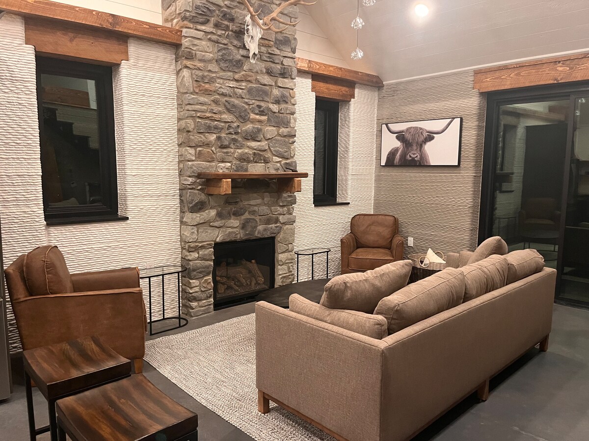A cozy living area is centered around a stone fireplace, framed by two large windows. Comfortable seating is provided by a spacious sofa and two armchairs. A wall artwork depicts a bull, adding a focal point to the neutral tones of the room.