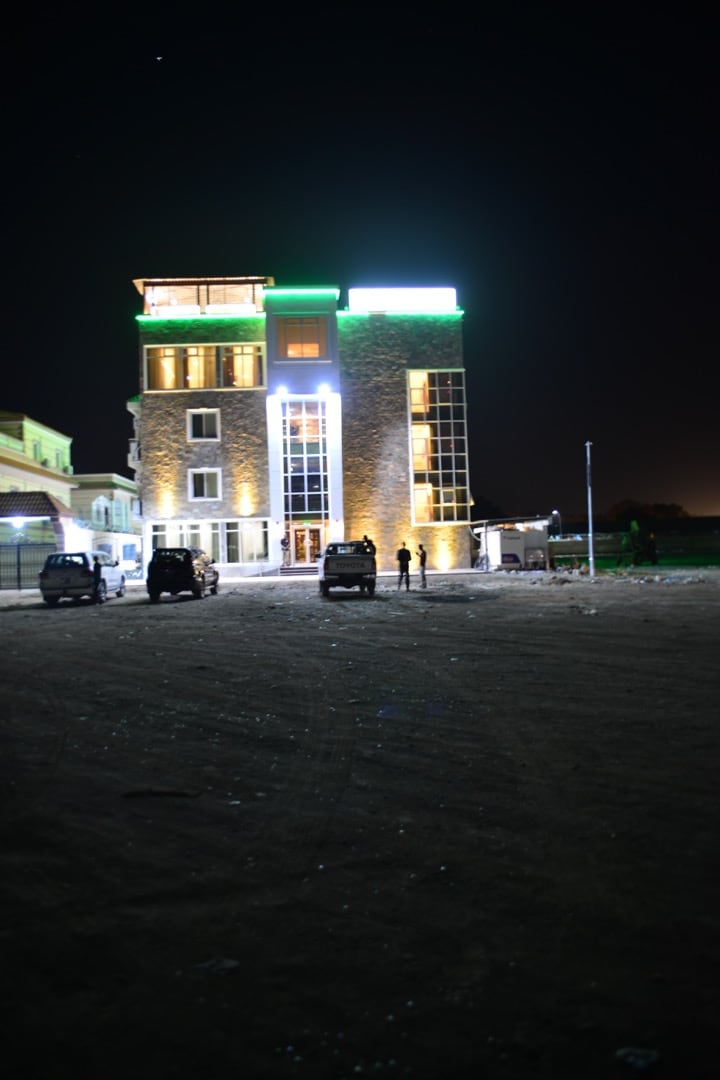 The exterior of a multi-story building is illuminated at night, highlighting its unique stone facade and large glass windows. Soft green and white lights accent the architectural features, creating a striking contrast against the dark sky.