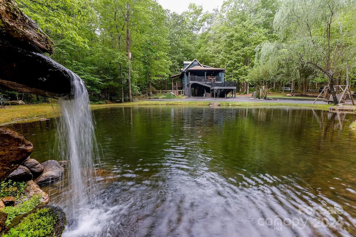 A serene setting is captured, featuring a tranquil pond with a gentle waterfall flowing into it. The cabin is visible in the background, surrounded by lush greenery, providing a peaceful retreat. The scene evokes a calm atmosphere, ideal for relaxation and connection with nature.