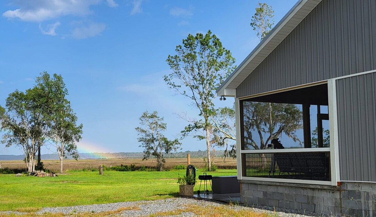 A cozy cabin exterior is framed by lush greenery, with large windows offering extensive views of the marshland. A rainbow is visible in the distance, complementing the serene landscape. A small patio area is also featured, enhancing the outdoor experience.