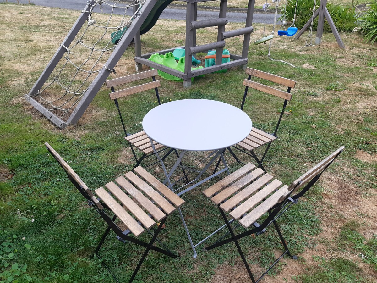A round white table is surrounded by four wooden folding chairs, set on a grassy area. In the background, a playground structure and green toys are visible, suggesting a space for outdoor relaxation and play.