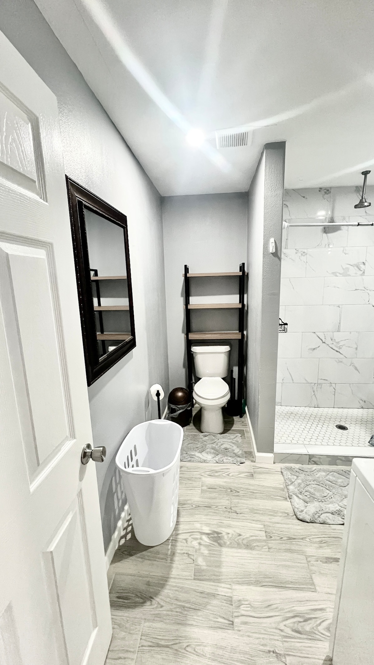 A modern bathroom featuring a shower with glass doors and marble tiles. A white toilet is positioned beside a slim shelving unit, while a mirror reflects light from the ceiling. A laundry basket is placed on the floor, accentuating the clean design.