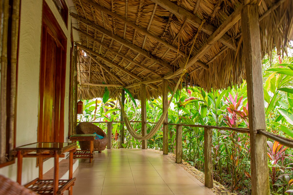 An outdoor terrace is displayed, featuring natural bamboo roofing and wooden railings. A hammock is suspended between the posts, with a wicker chair and small table positioned nearby. Lush greenery surrounds the area, enhancing the serene atmosphere.