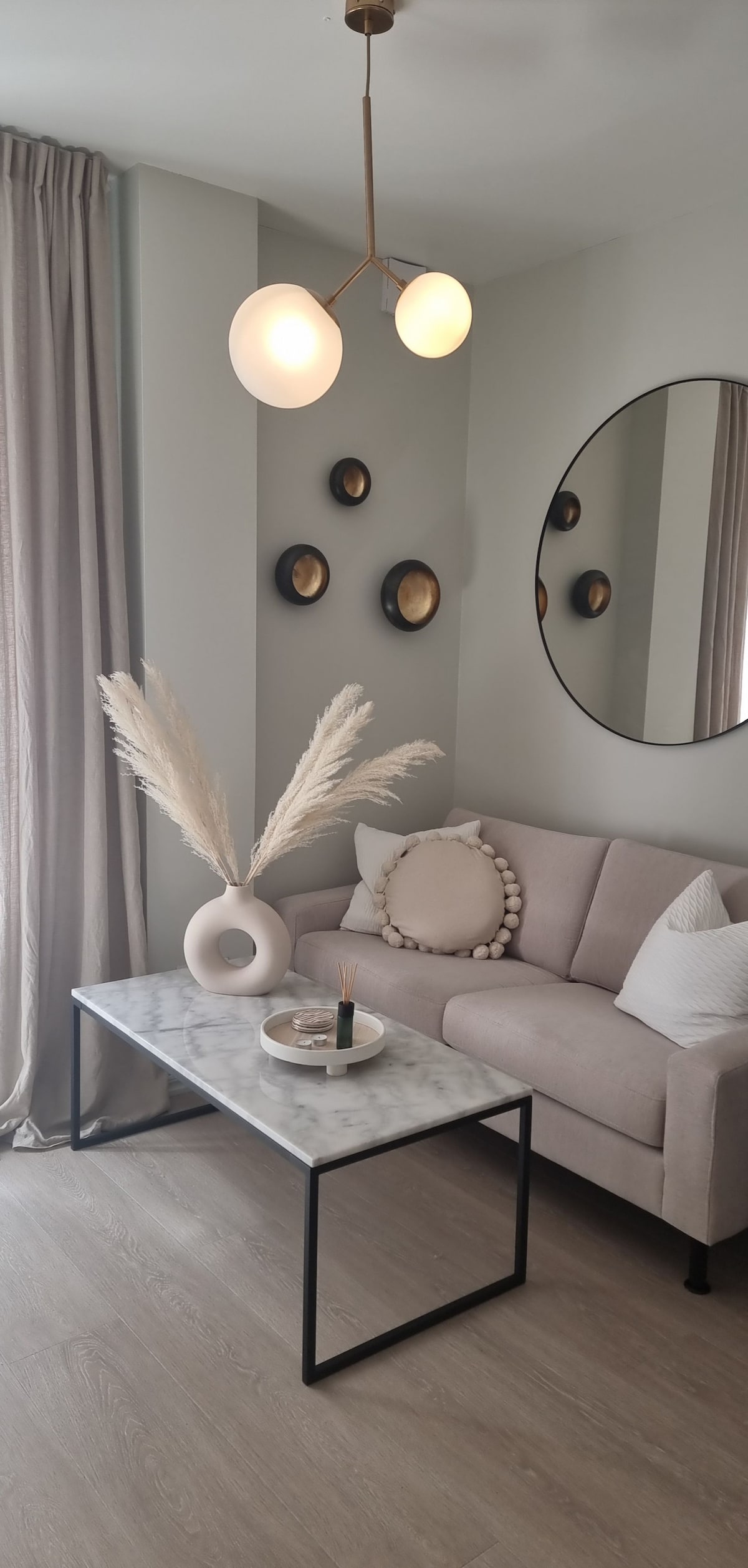 A light-filled living area features a soft gray sofa accented with decorative pillows. A round mirror reflects the space, while a marble coffee table holds a small vase and candle. Elegant wall decor and a stylish light fixture enhance the room's modern aesthetic.