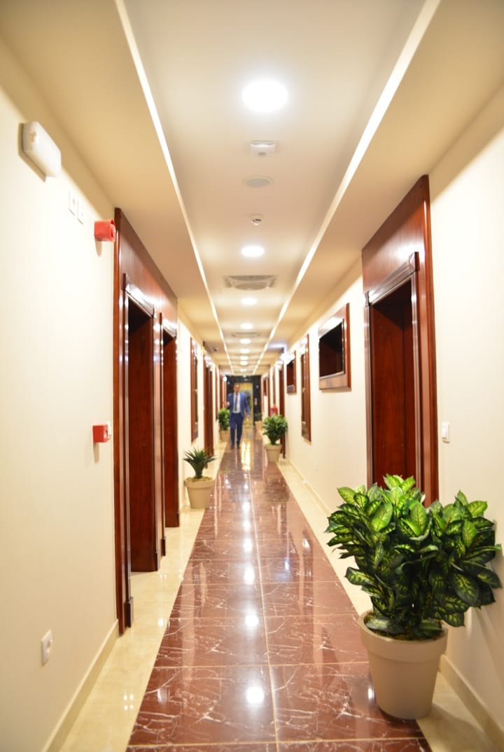 A long, bright hallway is lined with polished wooden doors leading to various rooms. The floor is finished with glossy tile, and potted plants are placed beside the walls, adding a touch of greenery to the setting. Soft lighting enhances the spacious feel of the corridor.