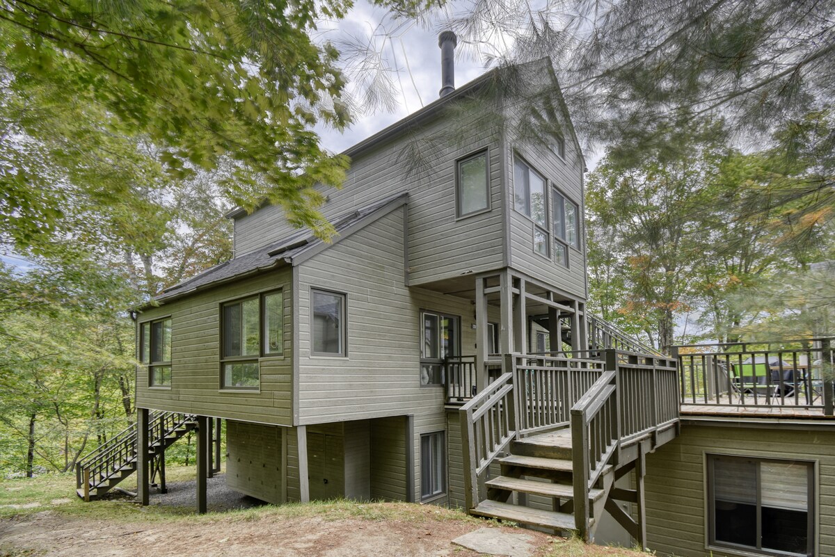A modern multi-level chalet is showcased, featuring a light green exterior and large windows that provide views of the surrounding trees. A deck extends from the building, complemented by a staircase leading to the ground level, creating a welcoming outdoor space.