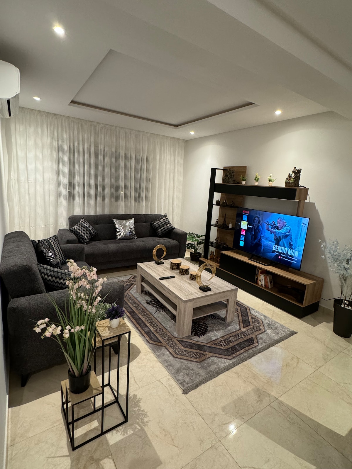 The living area features a modern seating arrangement with a dark sofa set and decorative cushions. A coffee table sits centrally, accompanied by a patterned rug. A TV stand displays a screen, while floral arrangements add a touch of elegance to the corner.