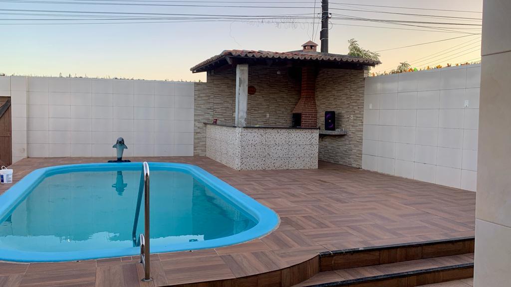 An outdoor area features a blue swimming pool surrounded by wooden decking. A stone barbecue area is visible nearby, complemented by a terracotta roof. The setting is enclosed by white walls, creating a private space with a hint of greenery in the background.