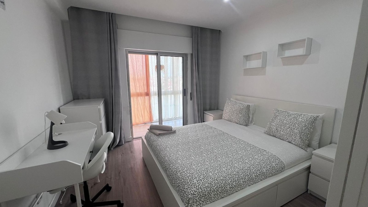 A bright bedroom is furnished with a spacious bed dressed in light bedding featuring a subtle pattern. A sleek desk and chair are positioned beside large windows, which are adorned with sheer curtains allowing soft natural light to filter through. Minimalist shelves are mounted on the walls.