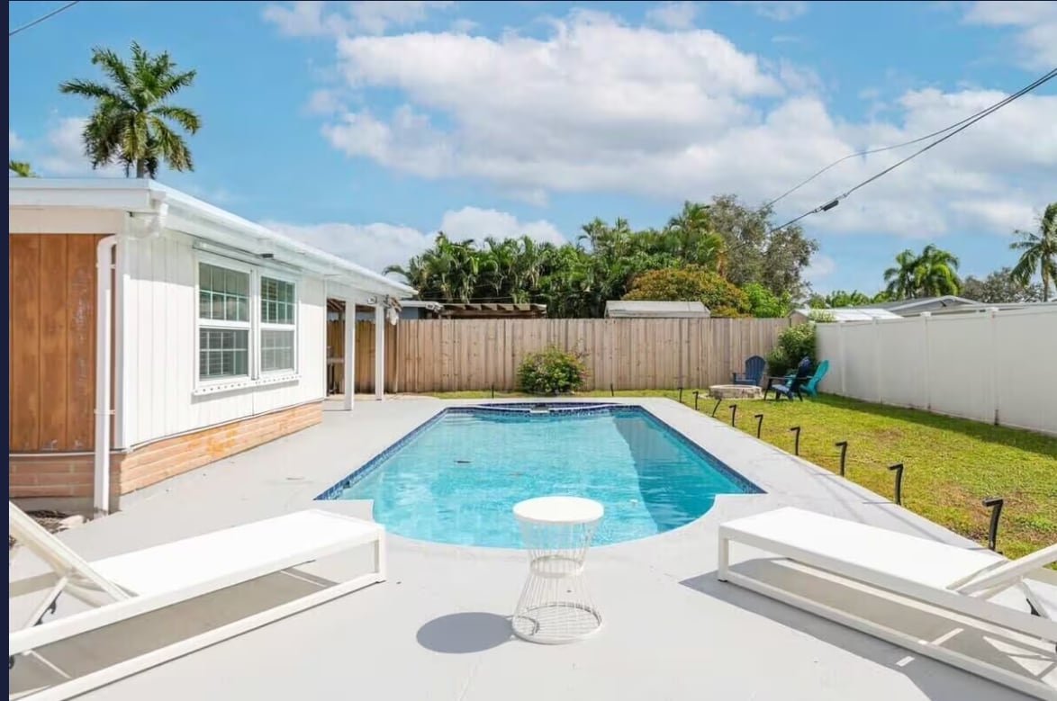 An inviting outdoor space features a large pool surrounded by a spacious deck. Two lounge chairs are positioned for relaxation, with greenery lining the perimeter. The home structure is visible in the background, complemented by a clear blue sky adorned with soft clouds.