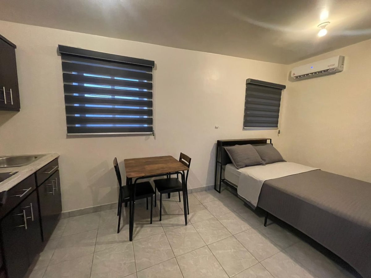 An inviting studio space features a comfortable bed paired with a wooden dining table and four chairs. A compact kitchen area is visible, along with modern window coverings that allow for controlled natural light. The air conditioning unit is positioned on the wall for optimal cooling.