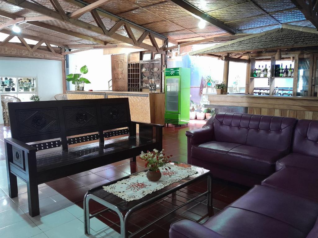 A communal lounge area is displayed, featuring a dark wooden bench and a purple sofa set. A coffee table sits in the center, adorned with a floral arrangement. The space is complemented by traditional decor elements and several indoor plants. A bar area is visible in the background.