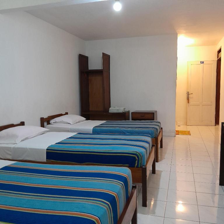 This neutral-toned room features three single beds, each with striped bed coverings. A wooden wardrobe stands along the wall, and a small drawer unit is positioned beside it. A bright door leads to the hallway, providing natural light to the room.