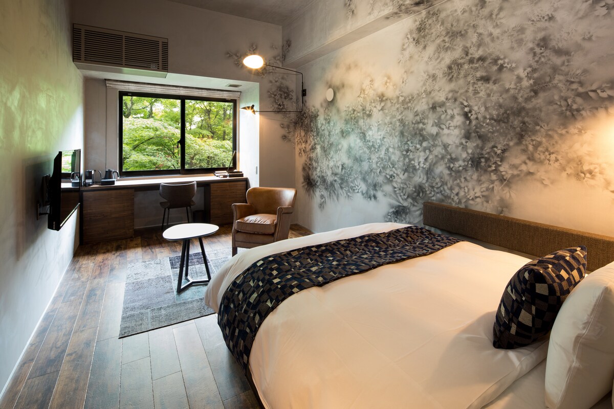 A single room features a spacious bed with textured bedding and decorative pillows. A wooden desk is positioned beside a large window, which frames views of the surrounding greenery. A comfortable armchair and a small round table are also present in the cozy space.