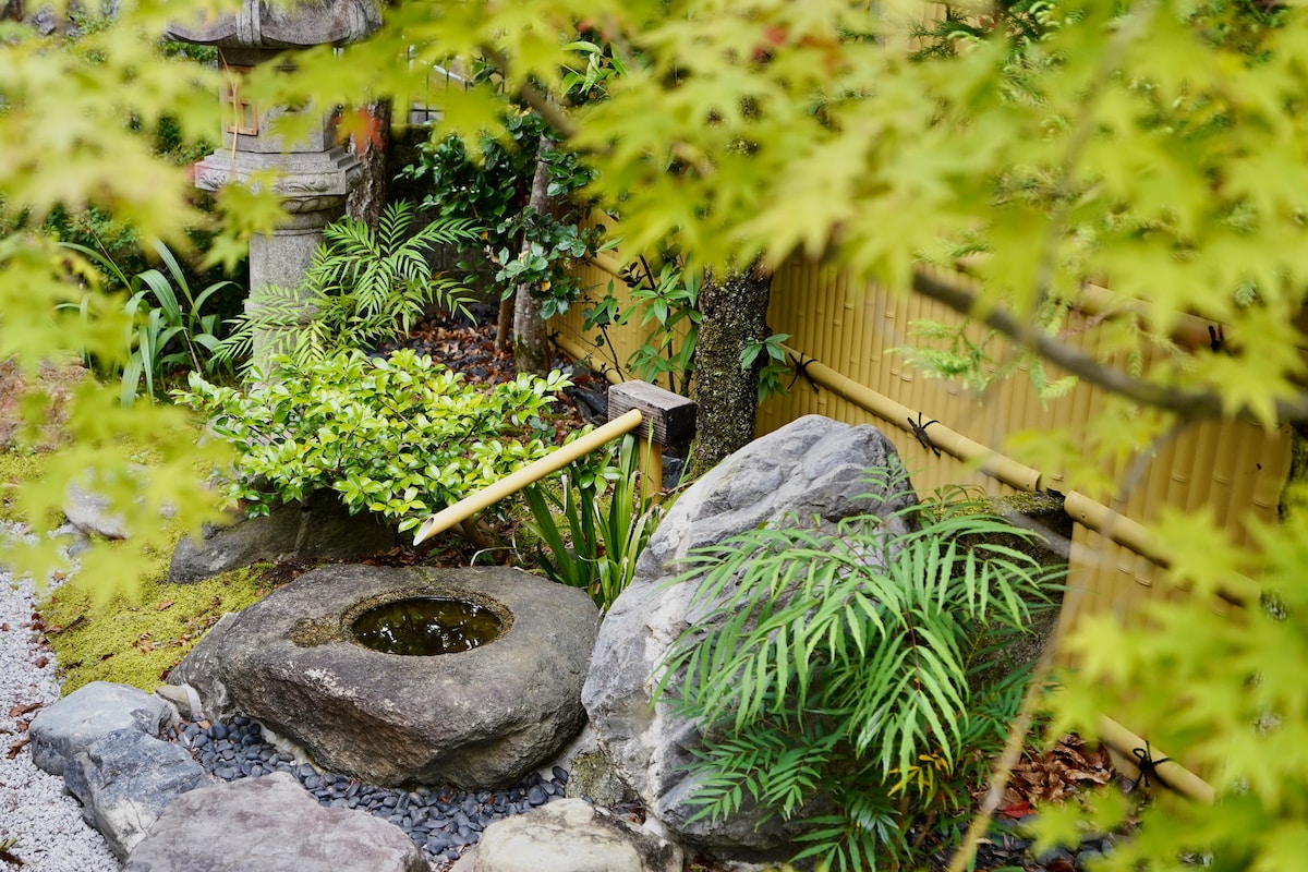 A serene Japanese garden showcases a stone basin nestled among lush greenery. Bamboo spouts gently flow water, surrounded by various plants and smooth stones, providing a tranquil atmosphere. A wooden fence serves as a backdrop, adding to the harmonious environment.