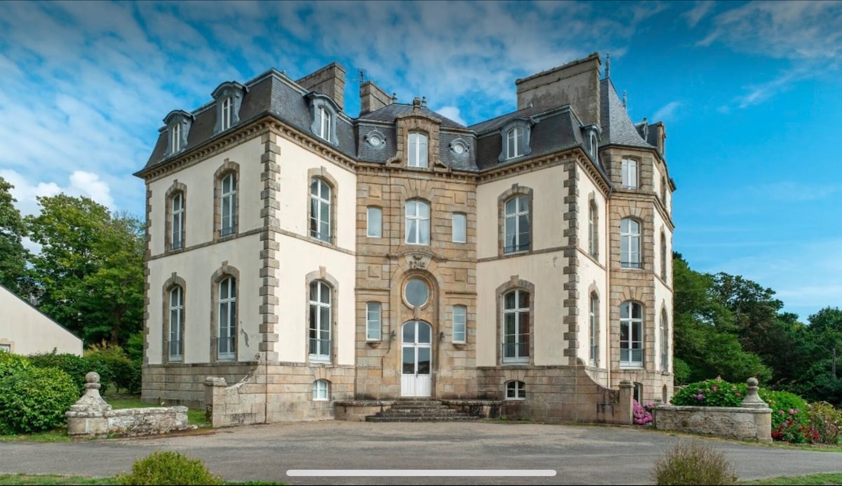 The historic castle exterior is showcased with elegant stone architecture and decorative features. Large arched windows and a grand entrance add to its charm. Lush greenery surrounds the building, complemented by colorful flower beds that enhance the serene outdoor space.