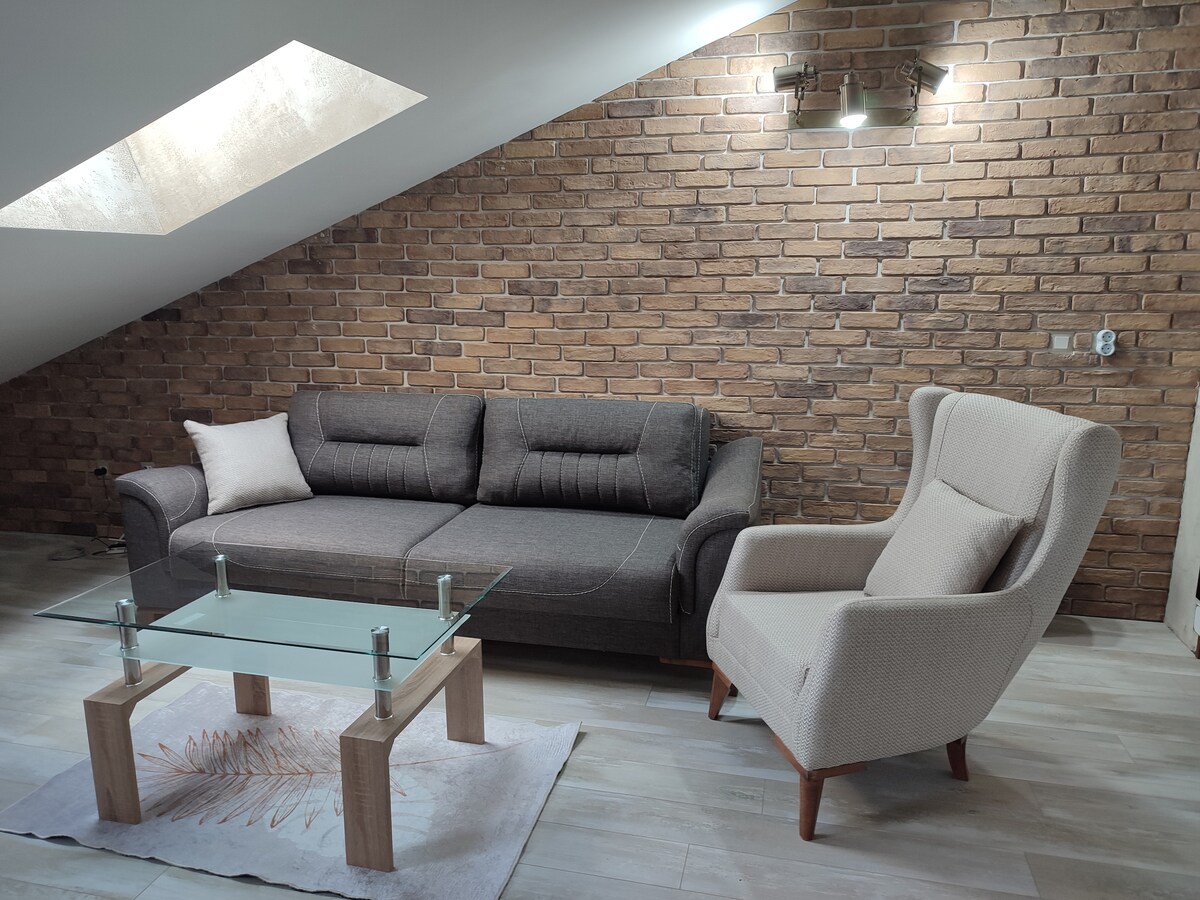 A comfortable living area features a dark sofa and a light armchair, positioned around a glass coffee table. The textured brick wall provides a rustic backdrop, while natural light enters through a skylight above.