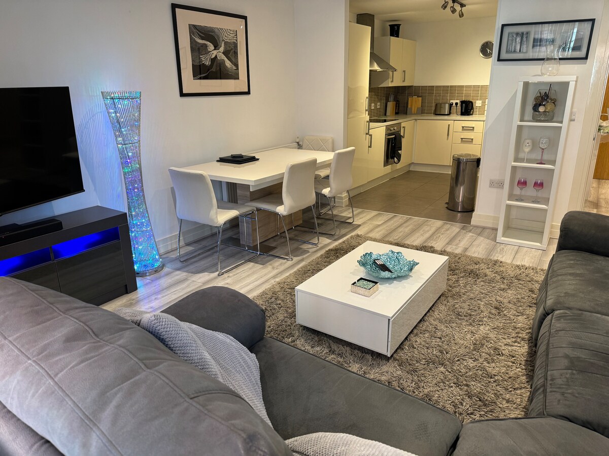 A spacious living area features a soft grey couch, offering a view of a sleek dining table with four chairs. A contemporary kitchen can be seen in the background, equipped with modern appliances. A lighted decorative column adds a vibrant touch, alongside a centerpiece displayed on the coffee table.