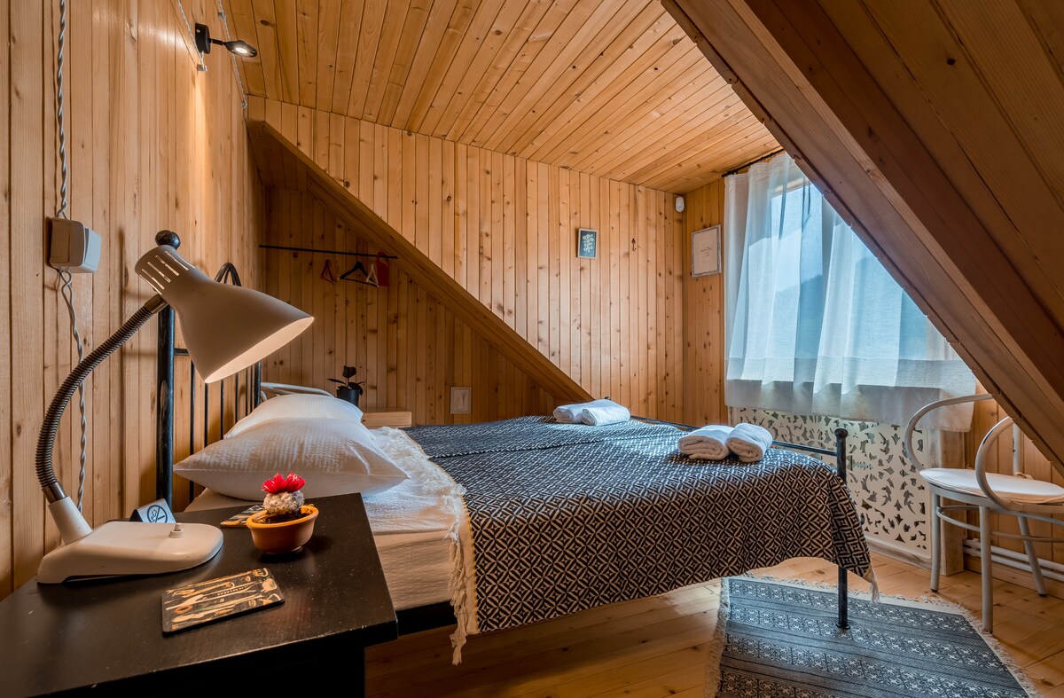 A wooden room features an angled ceiling with a light-colored wood paneling. A spacious bed with white linens and two neatly rolled towels is positioned beside a window adorned with light curtains. A bedside lamp and a small decorative bowl are placed on a dark table.