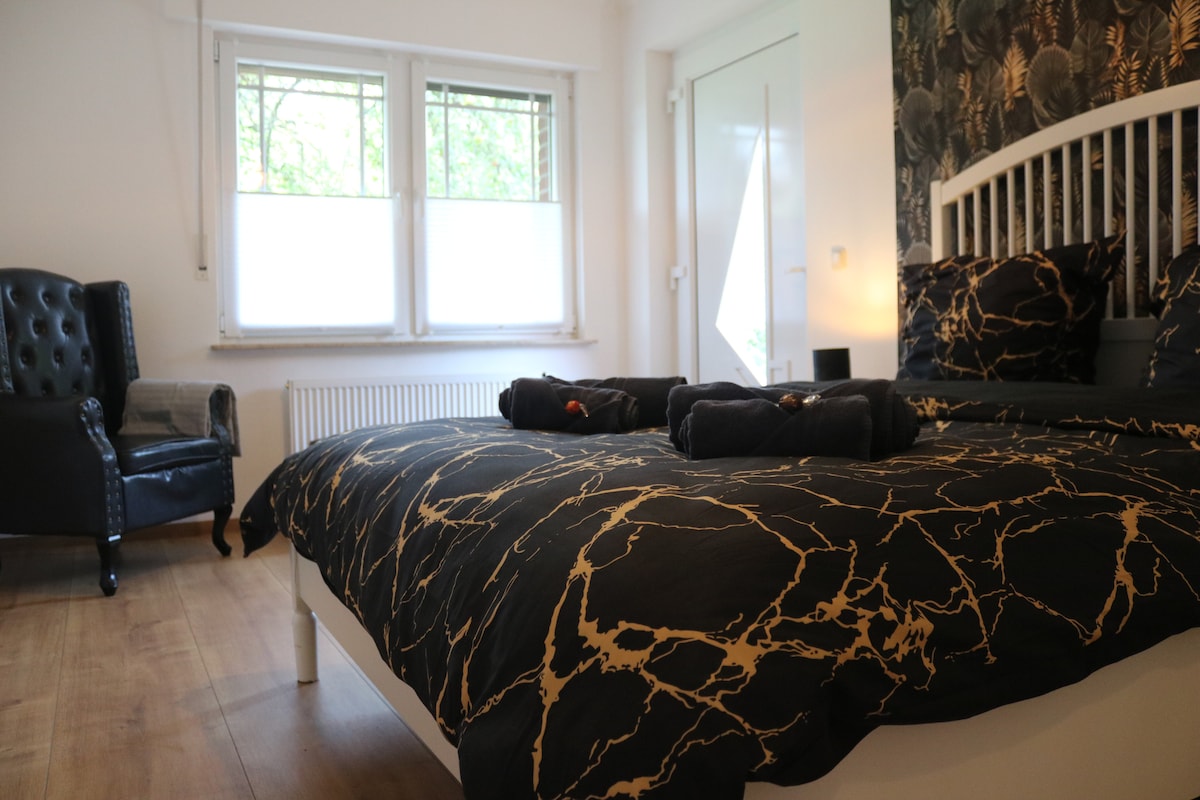 A sleeping area is presented with a spacious bed featuring dark bedding adorned with a marbled pattern. A comfortable chair is positioned in the corner, and natural light filters in through two windows, enhancing the room's bright and airy ambiance.