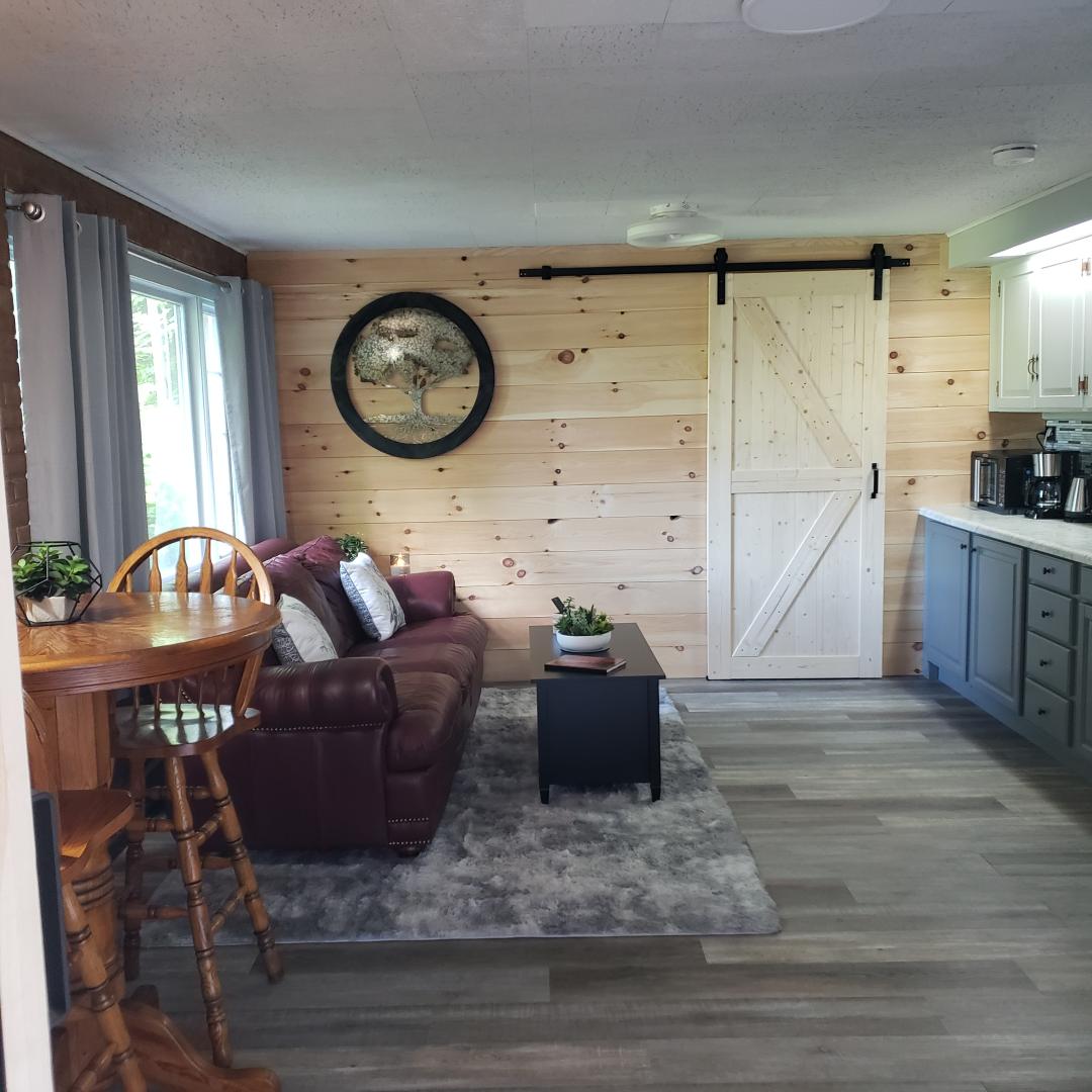 A living area features a soft leather couch paired with patterned throw pillows. A dark coffee table sits atop a plush rug. A small dining table with high chairs is positioned nearby. Light enters through large windows, complemented by light grey curtains.