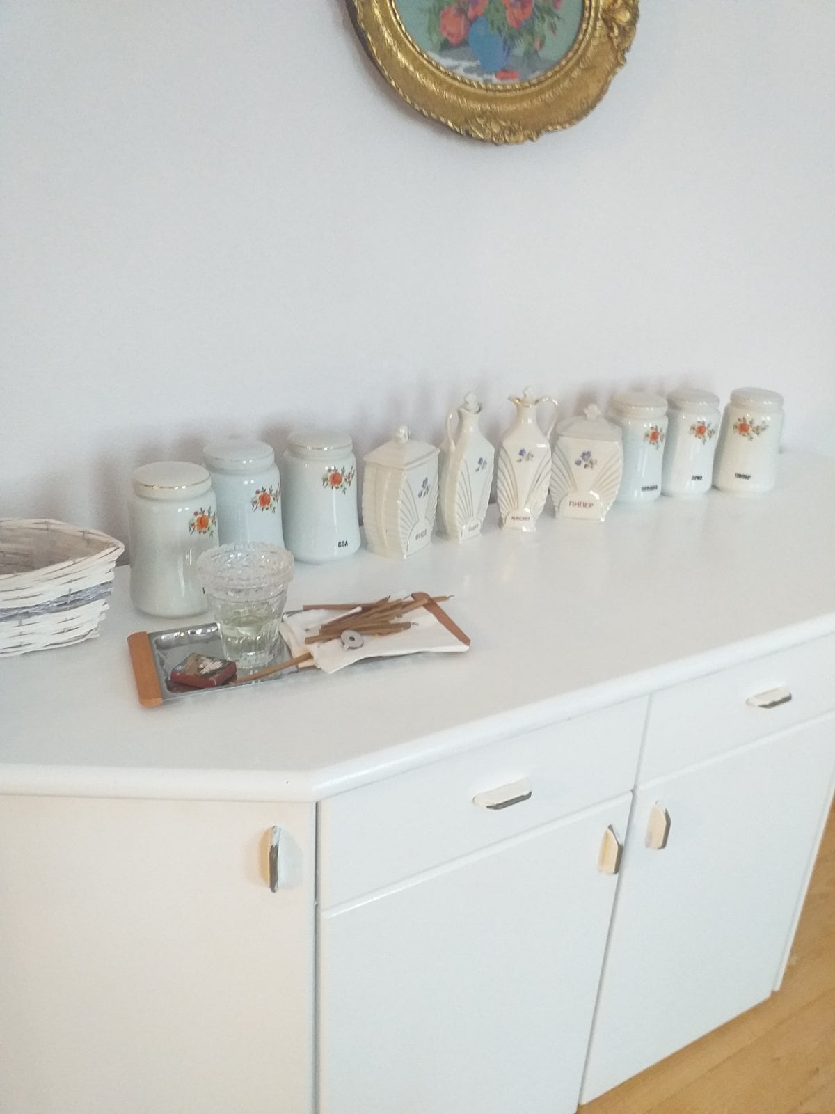 A white sideboard holds an array of ceramic containers, each featuring floral designs. A transparent glass, silver utensils, and a small basket are arranged neatly on top, contributing to a tidy and organized appearance.