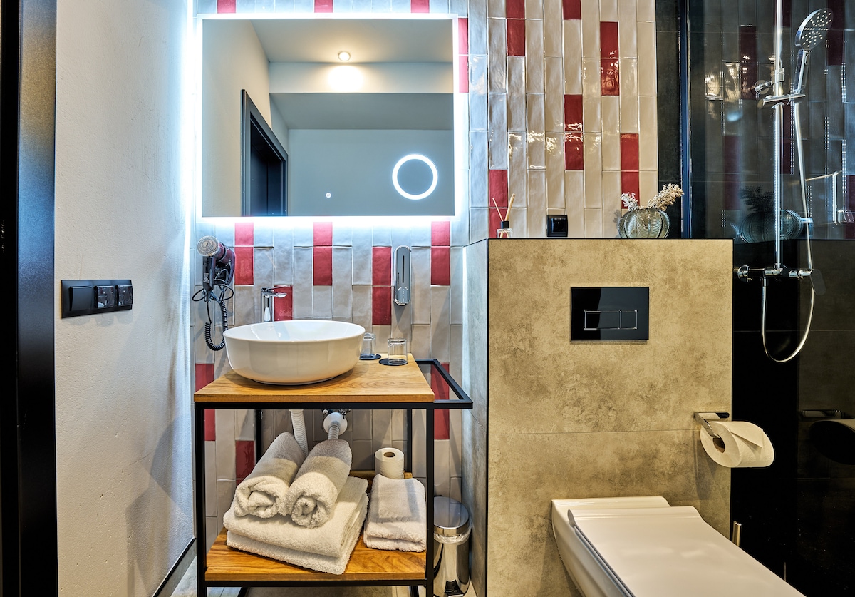 A modern bathroom features a stylish basin atop a wooden console, accompanied by neatly stacked towels. The illuminated mirror creates a bright ambiance. A shower area is visible in the background, showcasing contemporary tiles in red and white, while a sleek toilet is situated nearby.
