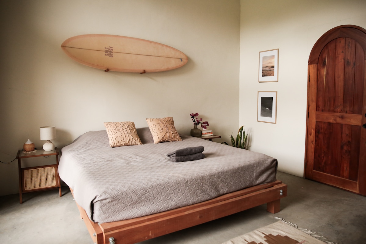 A spacious bedroom is presented with a large bed topped with neutral bedding and two decorative pillows. A surfboard is displayed on the wall above the bed, while a wooden door adds character. A small table, decorative plants, and framed artwork contribute to the room's aesthetic.